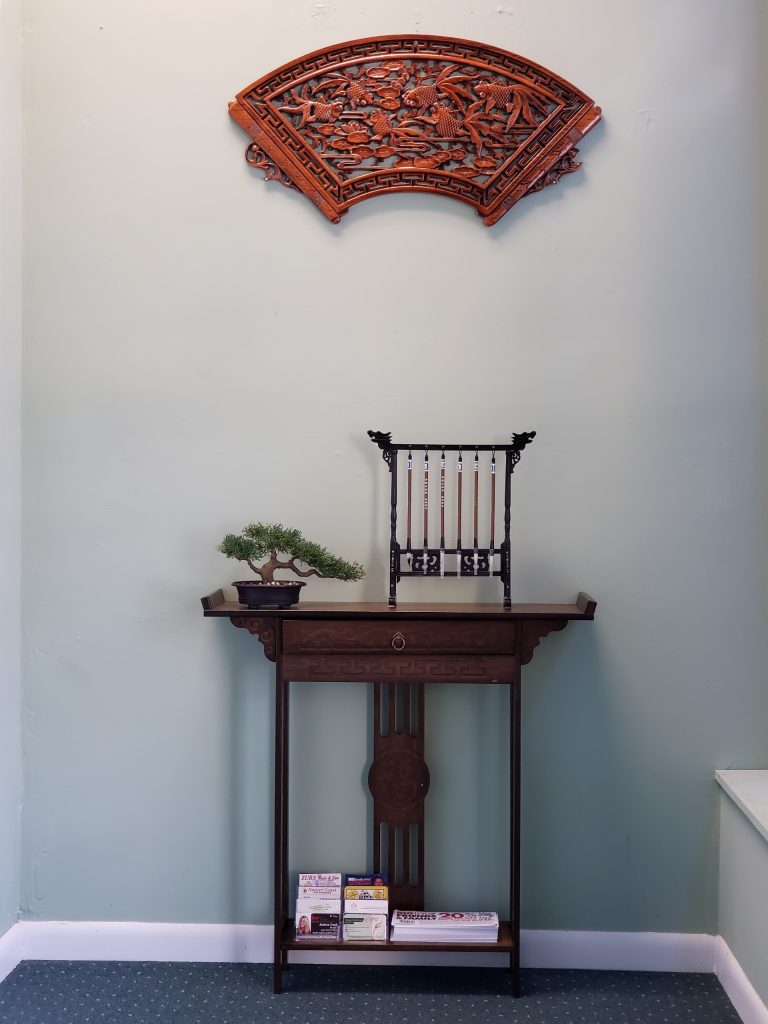 Dark wooden console table with a small bonsai on the left and a carved black rack on top, against a pale blue wall with a large red carved fan above.